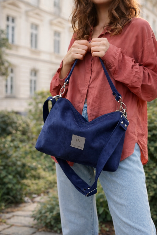 Mała zamszowa torebka w kolorze głębokiego granatu, elegancka i funkcjonalna. Miękka faktura, regulowany pasek i minimalistyczny design sprawiają, że idealnie pasuje do codziennych i eleganckich stylizacji.
