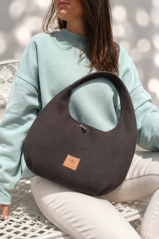 Zamszowa torebka hobo w ciemnobrązowym kolorze to połączenie minimalistycznego designu i funkcjonalności. Miękka faktura, pojemne wnętrze i wygodny pasek na ramię sprawiają, że idealnie sprawdzi się w codziennych stylizacjach casual i smart casual.